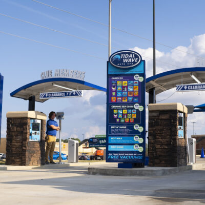 Tidal Wave Auto Spa entrance featuring various car wash options on a clear day, with a staff member ready to assist and signage promoting free vacuums with each wash.